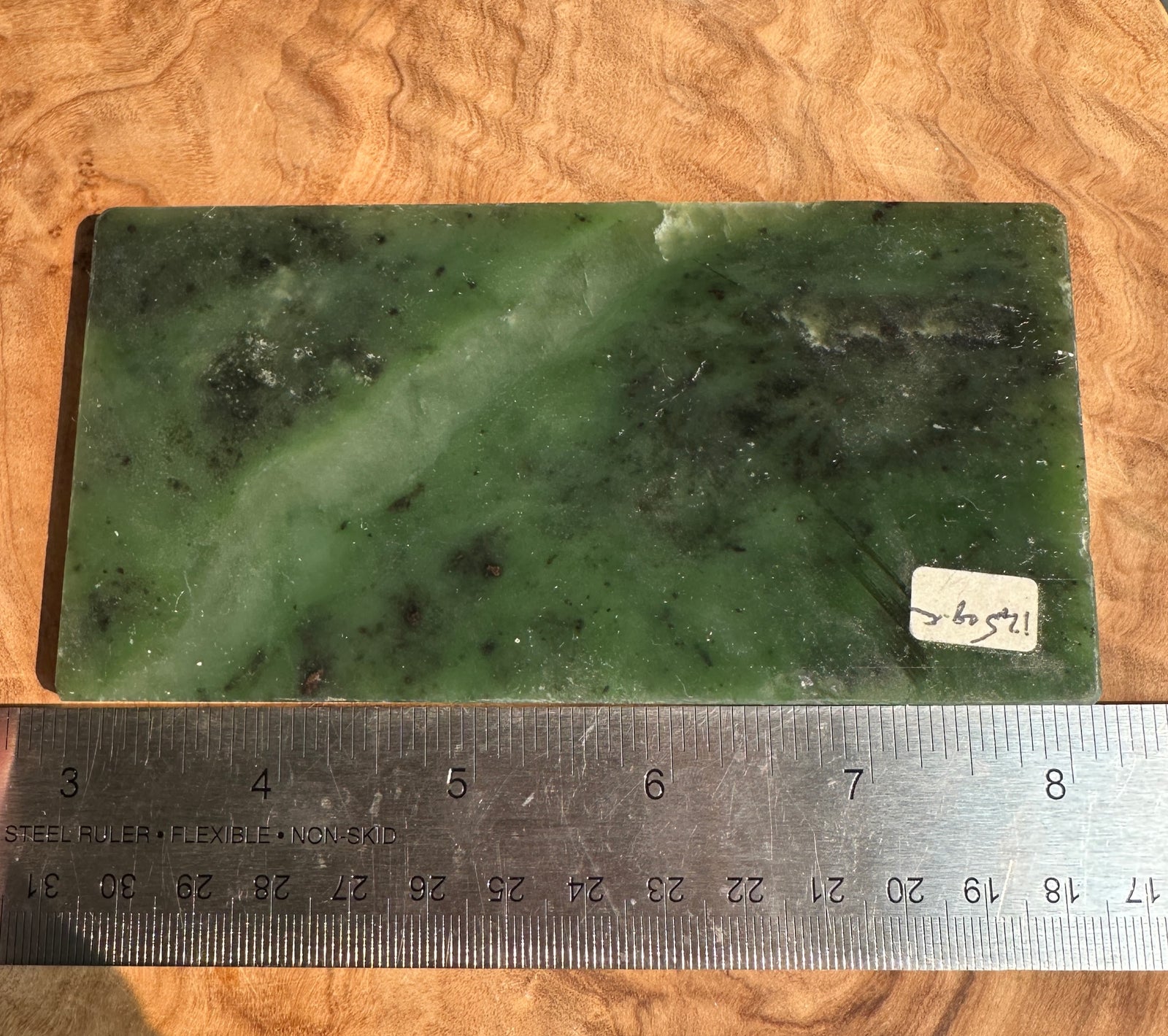 A rectangular green jade slab with natural marbling and dark inclusions, placed on a wood-grain surface with a metal ruler along the bottom edge for scale.