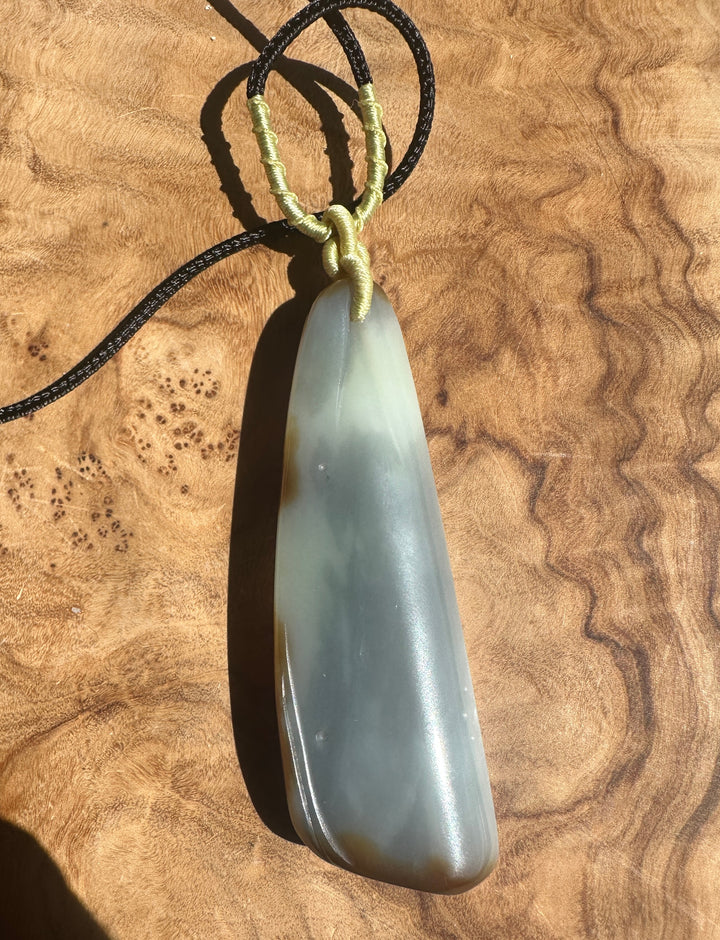 Pale jade Loulan pendant with a brown stripe, suspended by a wrapped beige cord over a wooden surface