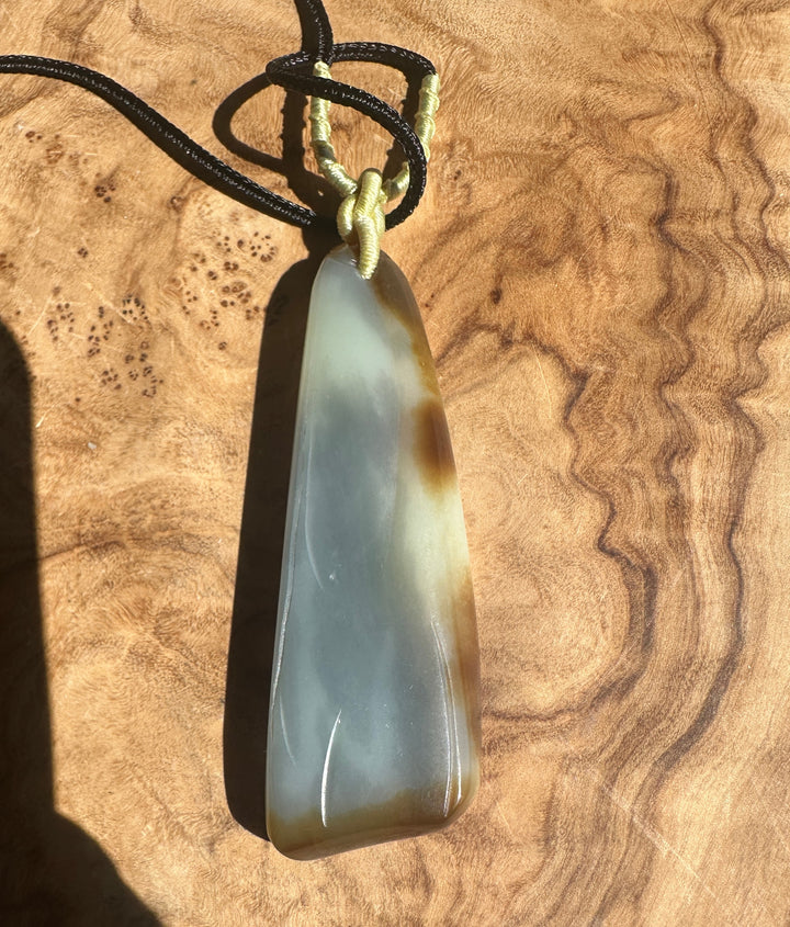 Pale green nephrite jade pendant with brown streaks hanging from a black cord with beige wrap on a wood background