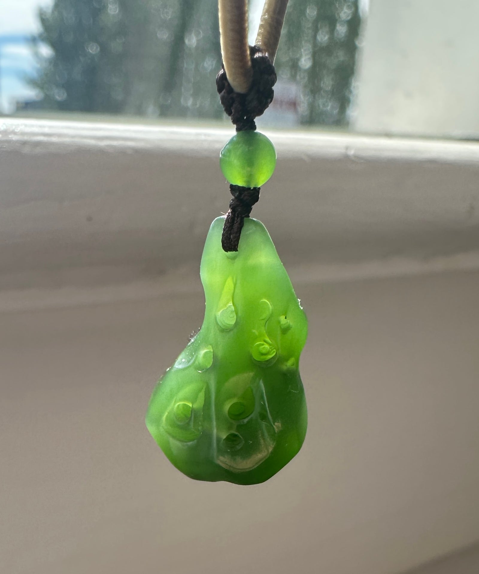 Green round bead and carved pendant on a dark braided cord against a window background