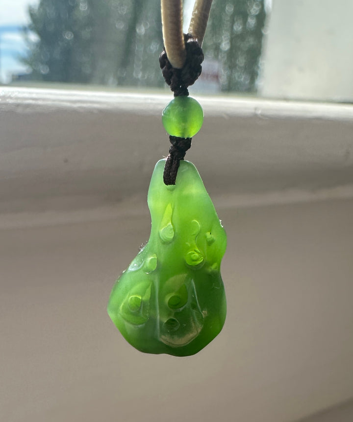 Green round bead and carved pendant on a dark braided cord against a window background