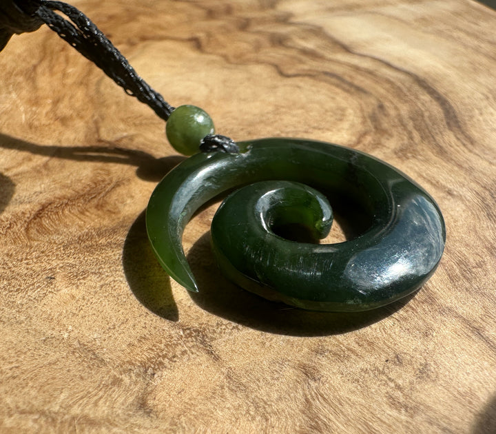 A dark green polished stone spiral pendant necklace with a matching round bead and black cord, resting on a textured wooden surface.