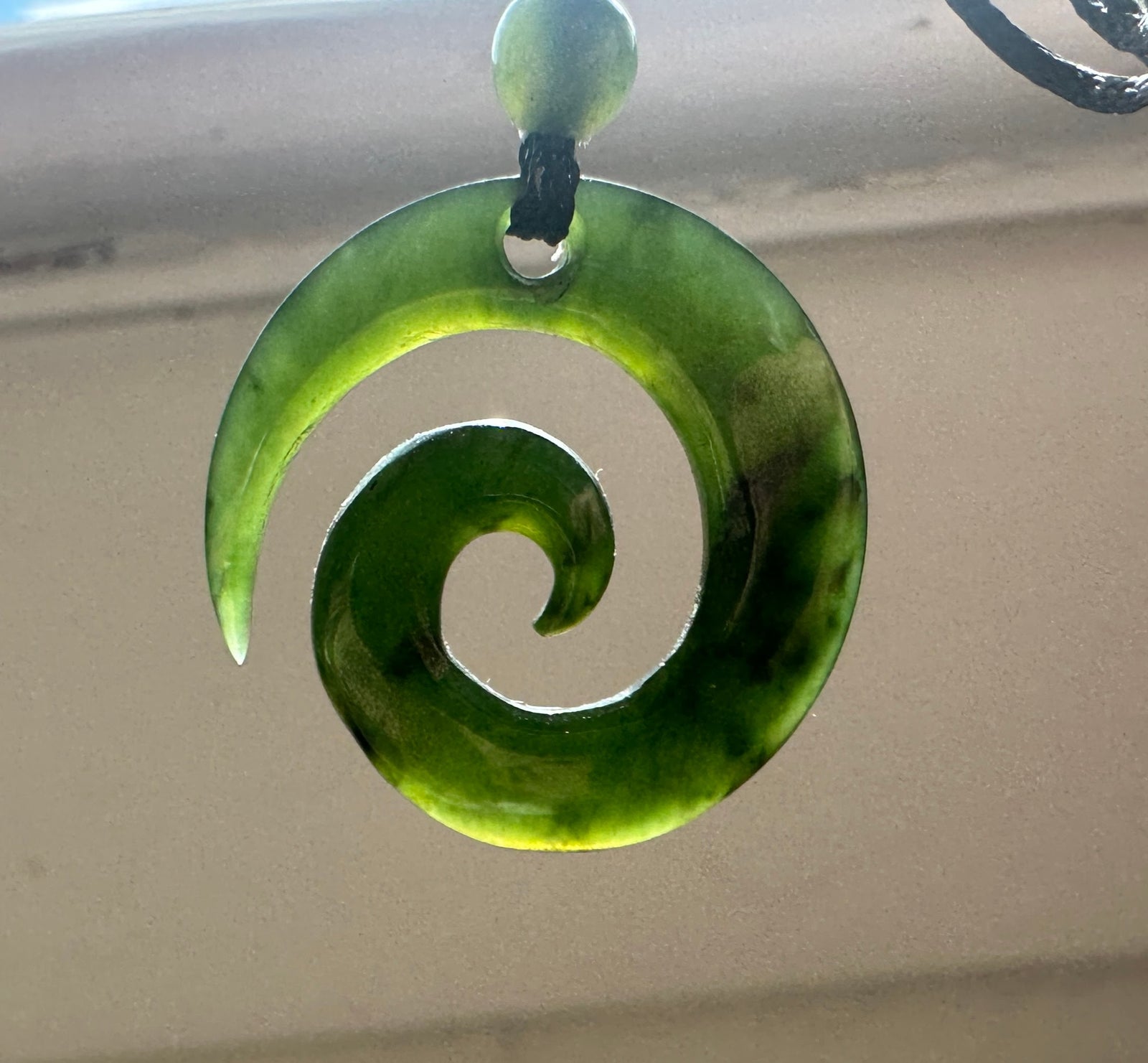 A green carved spiral pendant, hanging from a black cord with a small green bead. The pendant is photographed against a soft-focus beige background.