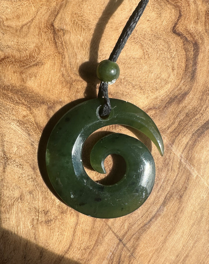 A dark green carved stone pendant necklace with a spiral design, hanging from a black cord with a small green bead, displayed on a textured wooden surface.