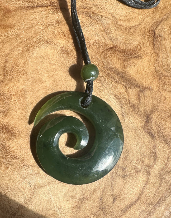 A dark green carved jade pendant necklace with a spiral design, hanging from a black cord with a small green bead, set against a textured wood background.