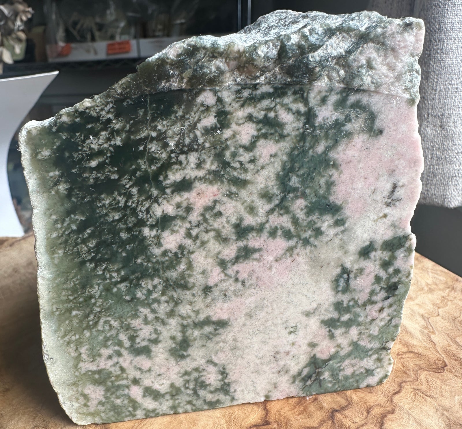 A raw, rough-cut stone slab with a marbled pattern of dark green, pale green, and pinkish-beige, standing upright on a wooden surface.