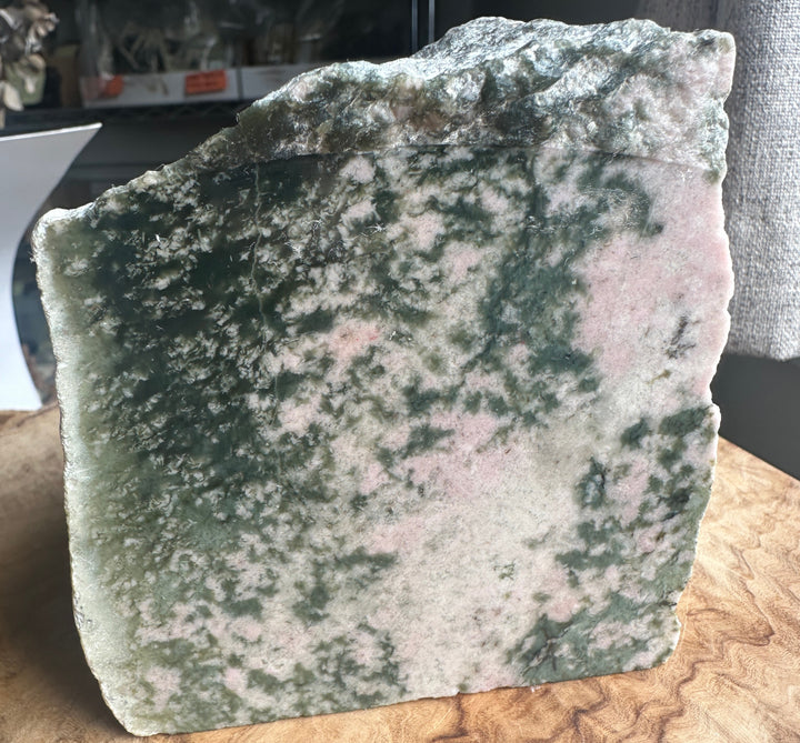 A raw, rough-cut stone slab with a marbled pattern of dark green, pale green, and pinkish-beige, standing upright on a wooden surface.