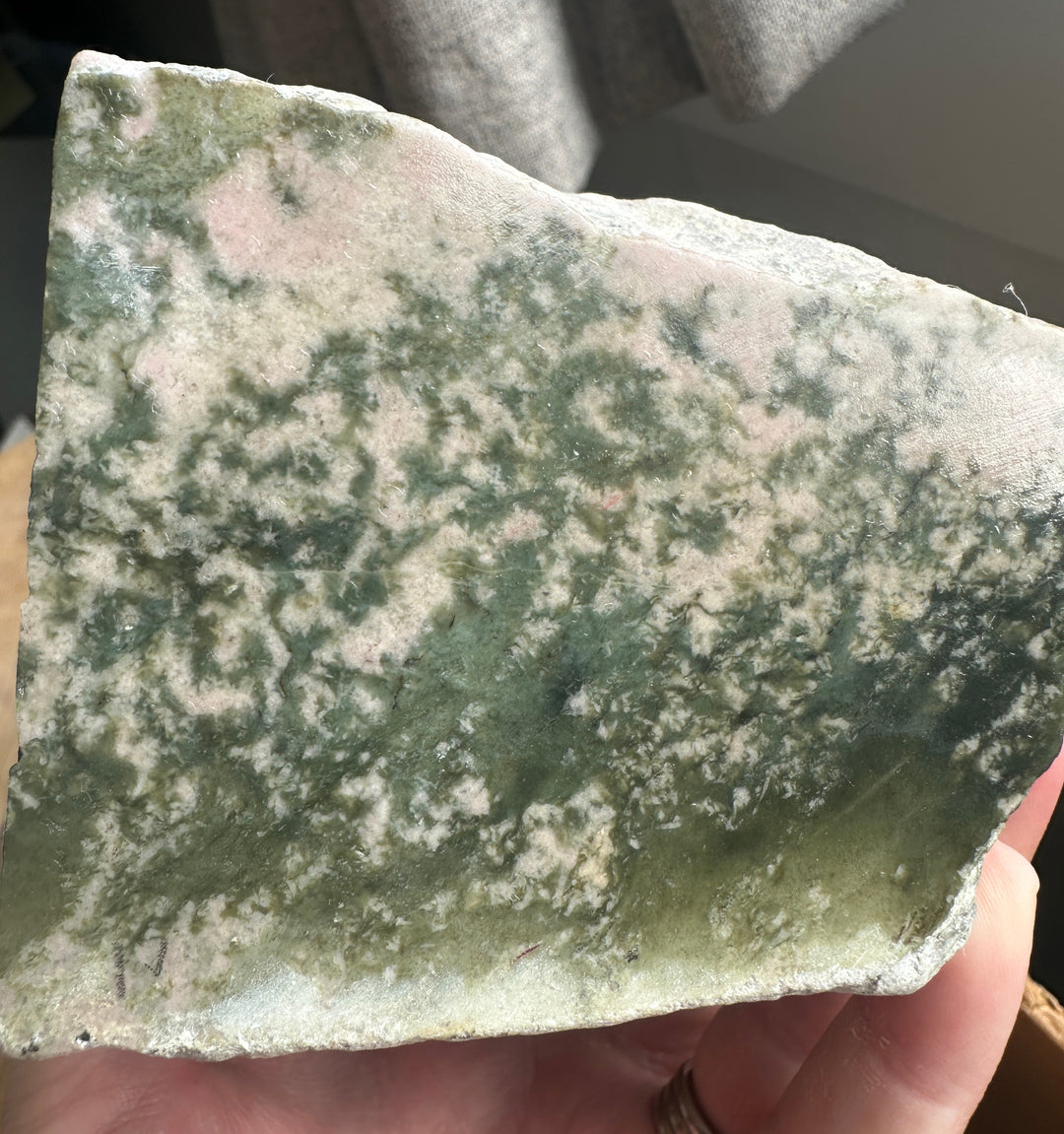 A hand holding a raw, rough stone slab with a mottled pattern of green, beige, and light pink, photographed in a casual indoor setting.
