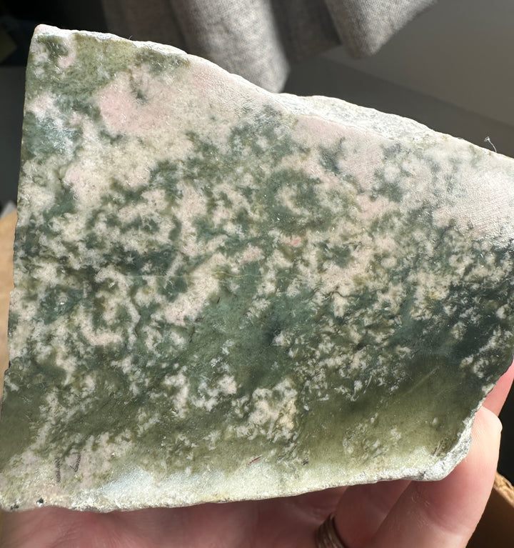 A hand holding a raw, rough stone slab with a mottled pattern of green, beige, and light pink, photographed in a casual indoor setting.