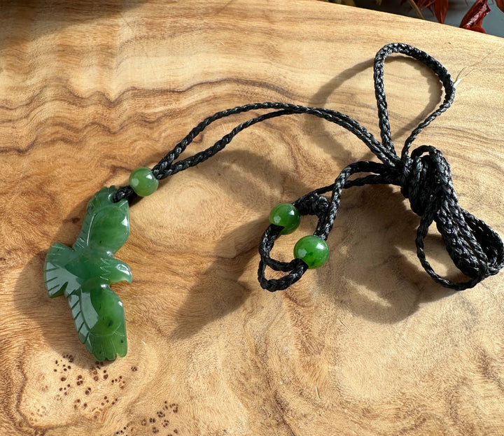 Jade eagle pendant on a braided black cord with green beads, displayed on a wooden surface.