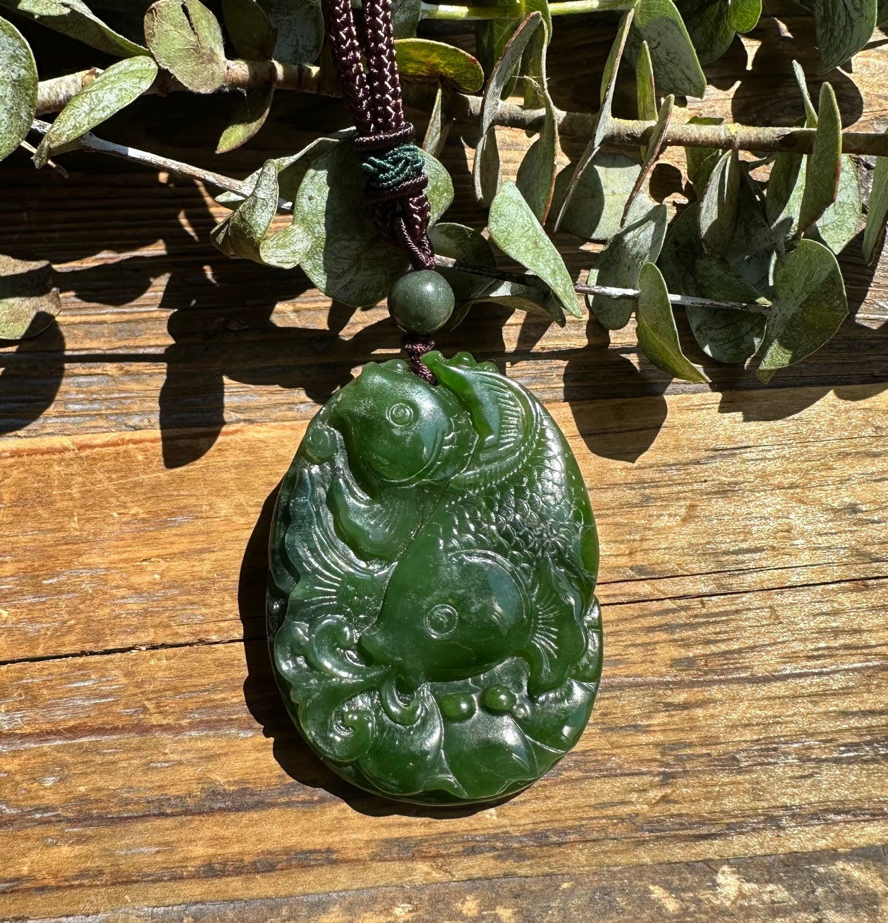Green jade koi fish pendant in a rounded oval form with two fish and swirling water details on a dark braided cord
