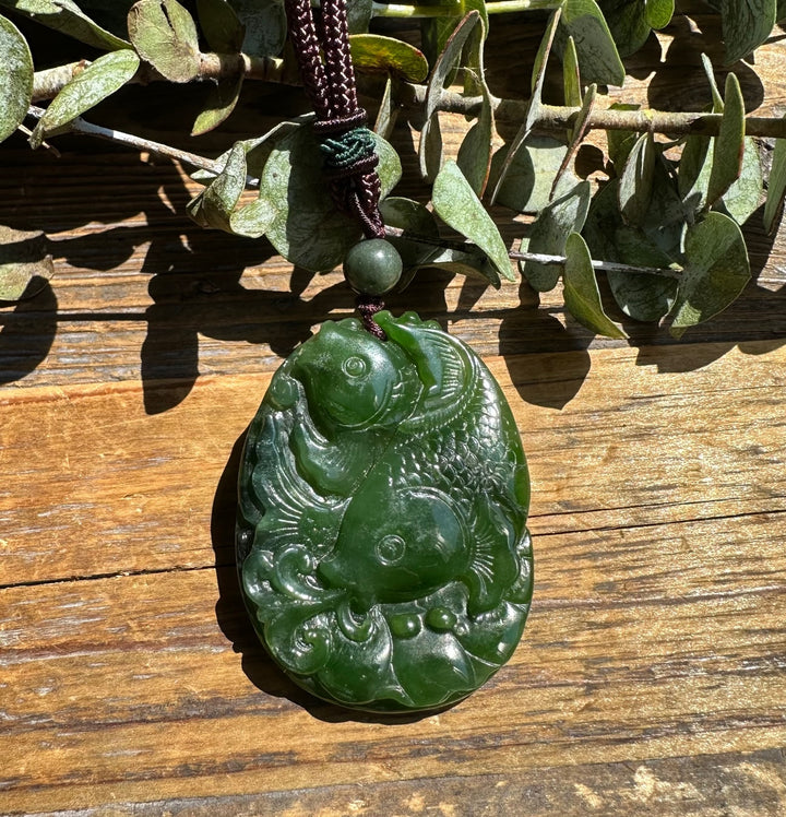 Green jade koi fish pendant in a rounded oval form with two fish and swirling water details on a dark braided cord