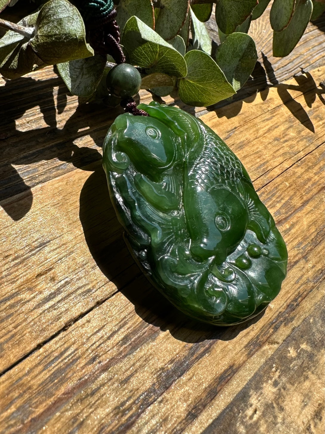 Close up of a green jade koi fish pendant with detailed carving and flowing patterns suspended from a dark cord