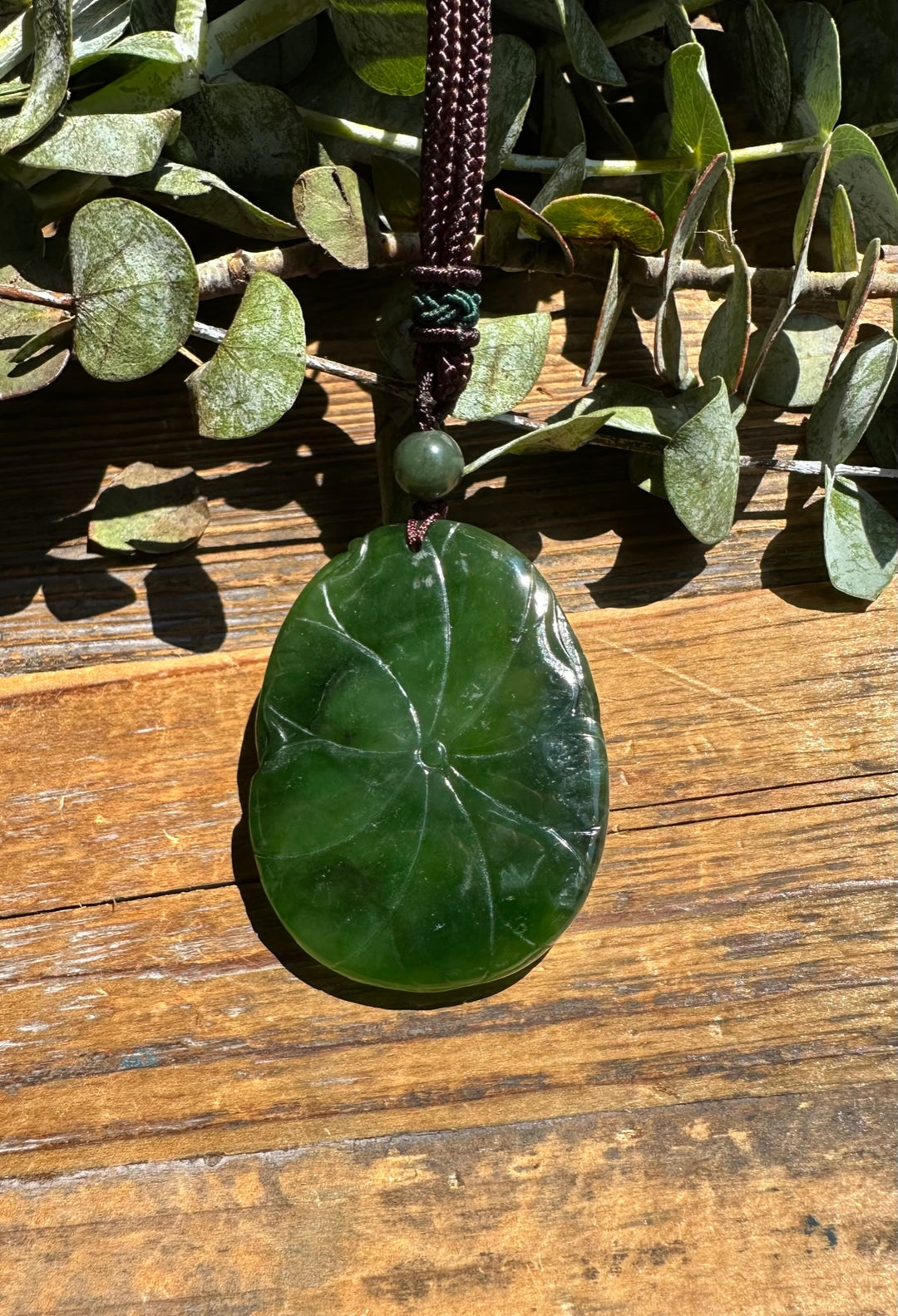 Deep green round jade pendant with natural swirl patterns hanging from a brown cord with a green bead