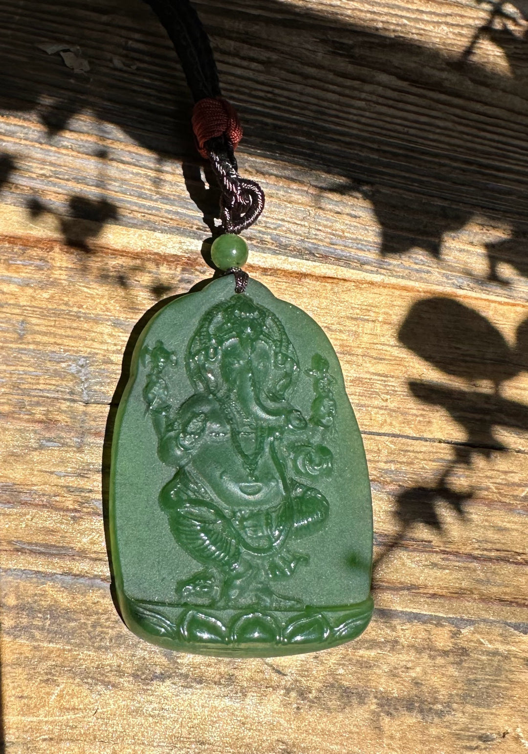 Close-up of green jade Ganesha pendant showing intricate relief on a rounded tablet, attached to a braided cord with a green bead.