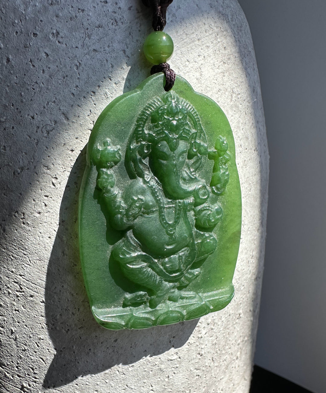 Green jade Ganesha Buddha pendant carved in relief on a rounded plaque, hanging from a brown cord with a green bead.