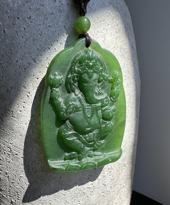 Green jade Ganesha Buddha pendant carved in relief on a rounded plaque, hanging from a brown cord with a green bead.