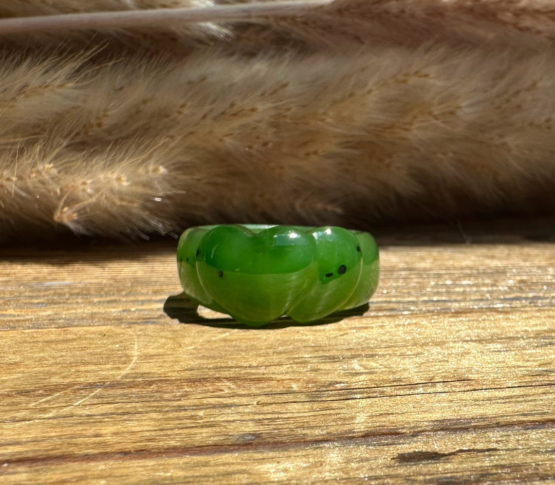 A carved green jade ring with a smooth, rounded, heart shape, resting on a rustic wooden surface with dried grass in the background.