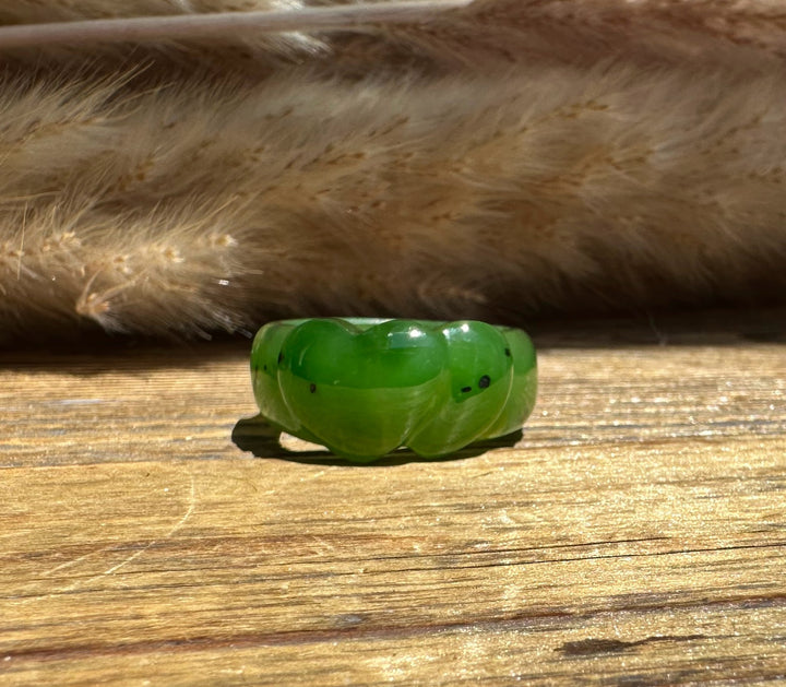 A carved green jade ring with a smooth, rounded, heart shape, resting on a rustic wooden surface with dried grass in the background.