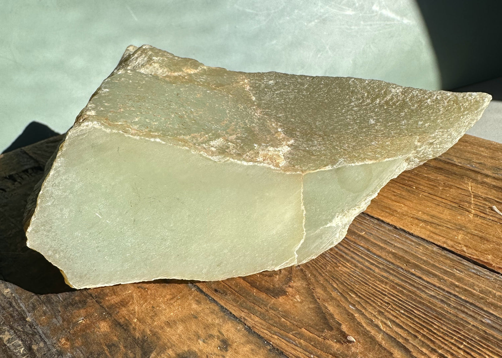Korean Jade Rough chunk with pale green surface and brown crust on top resting on a wooden table