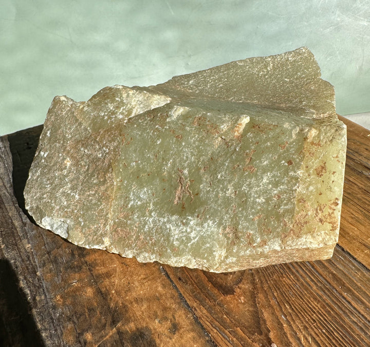 Korean Jade Rough block with green gray surface and rust colored specks captured from a side angle on wood