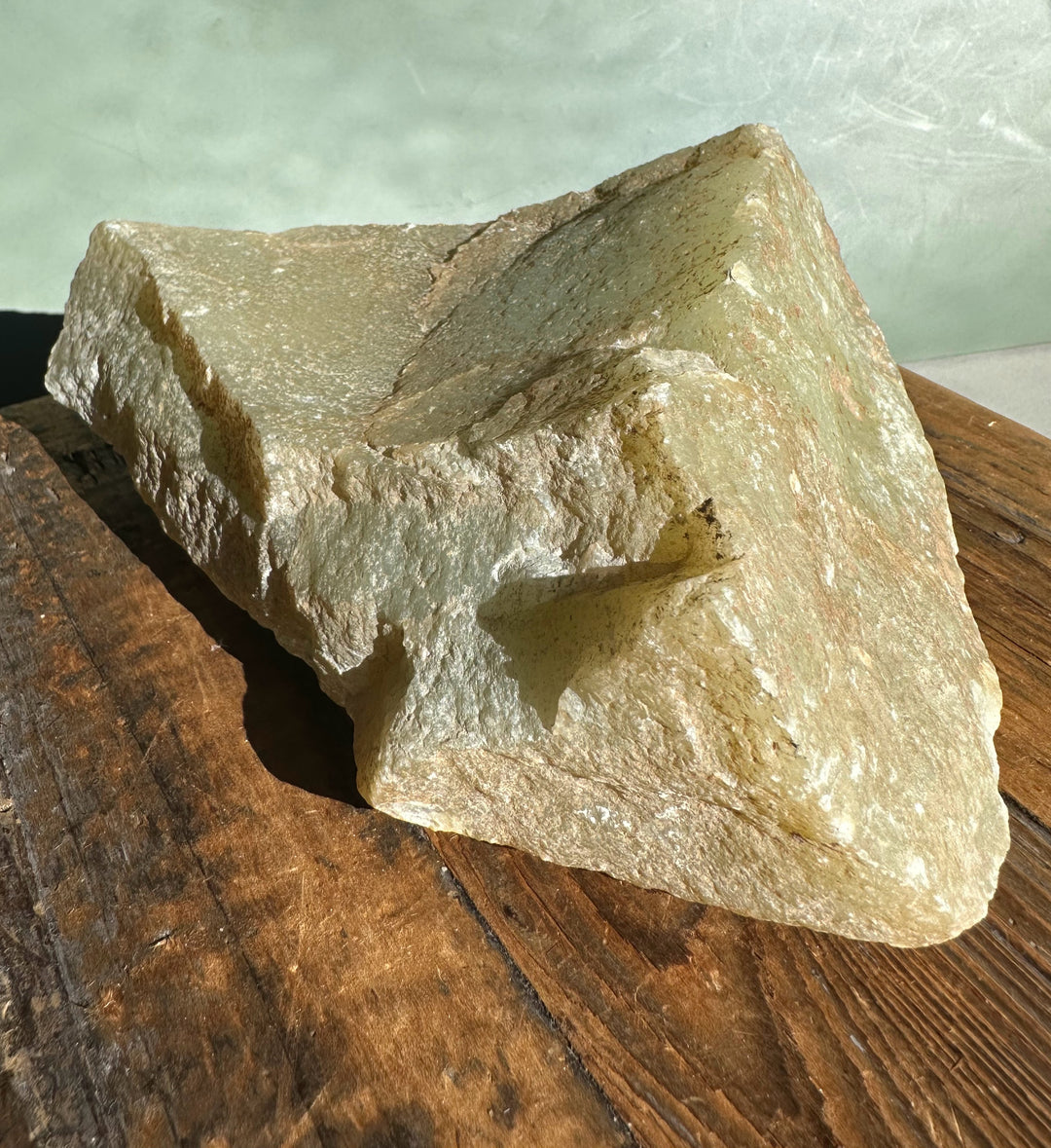 Rough Korean jade specimen with pale green beige surface on a wooden table.