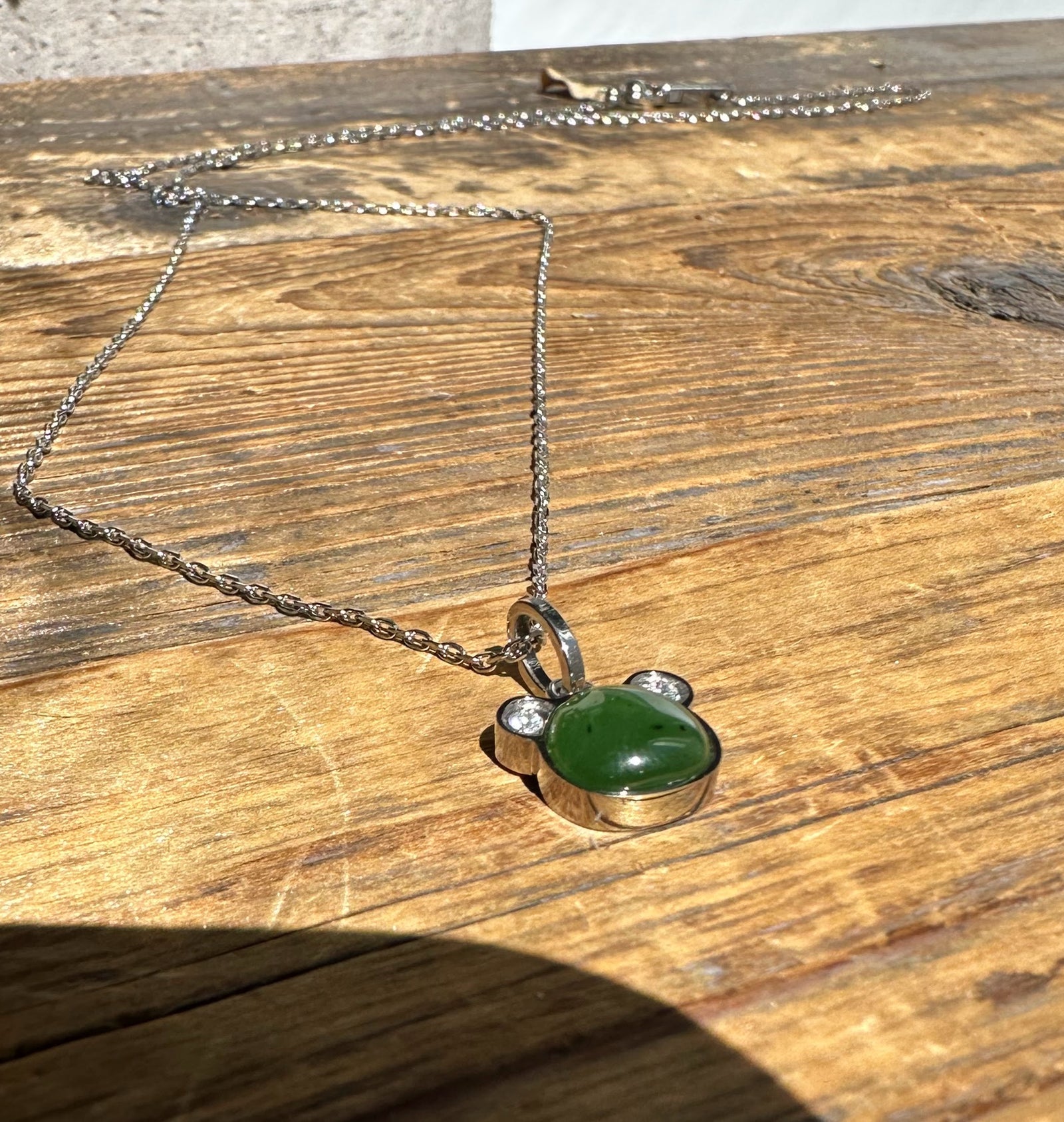 Green nephrite jade teddy bear pendant on a silver chain resting on sunlit wooden surface