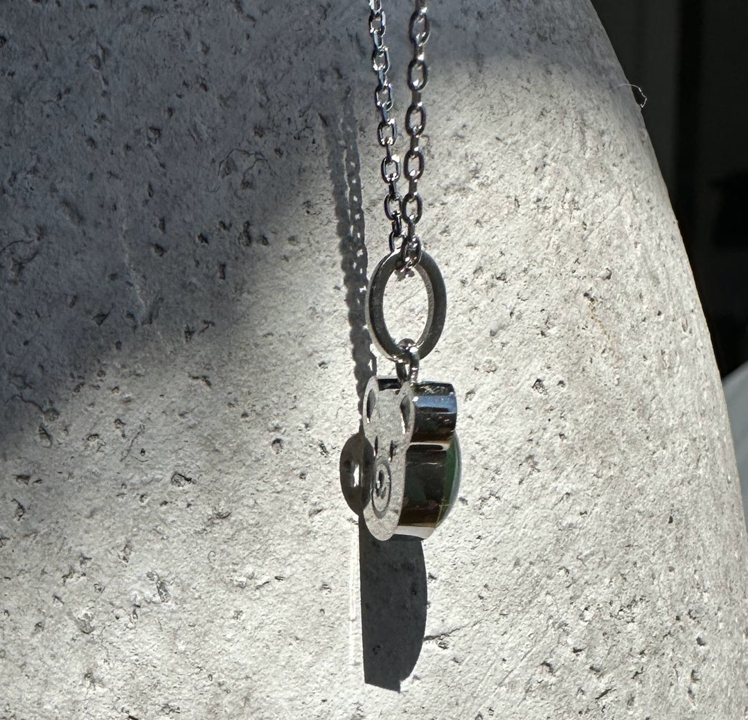 Side view of the teddy bear pendant with circular metal charms hanging from a silver chain against a pale stone background