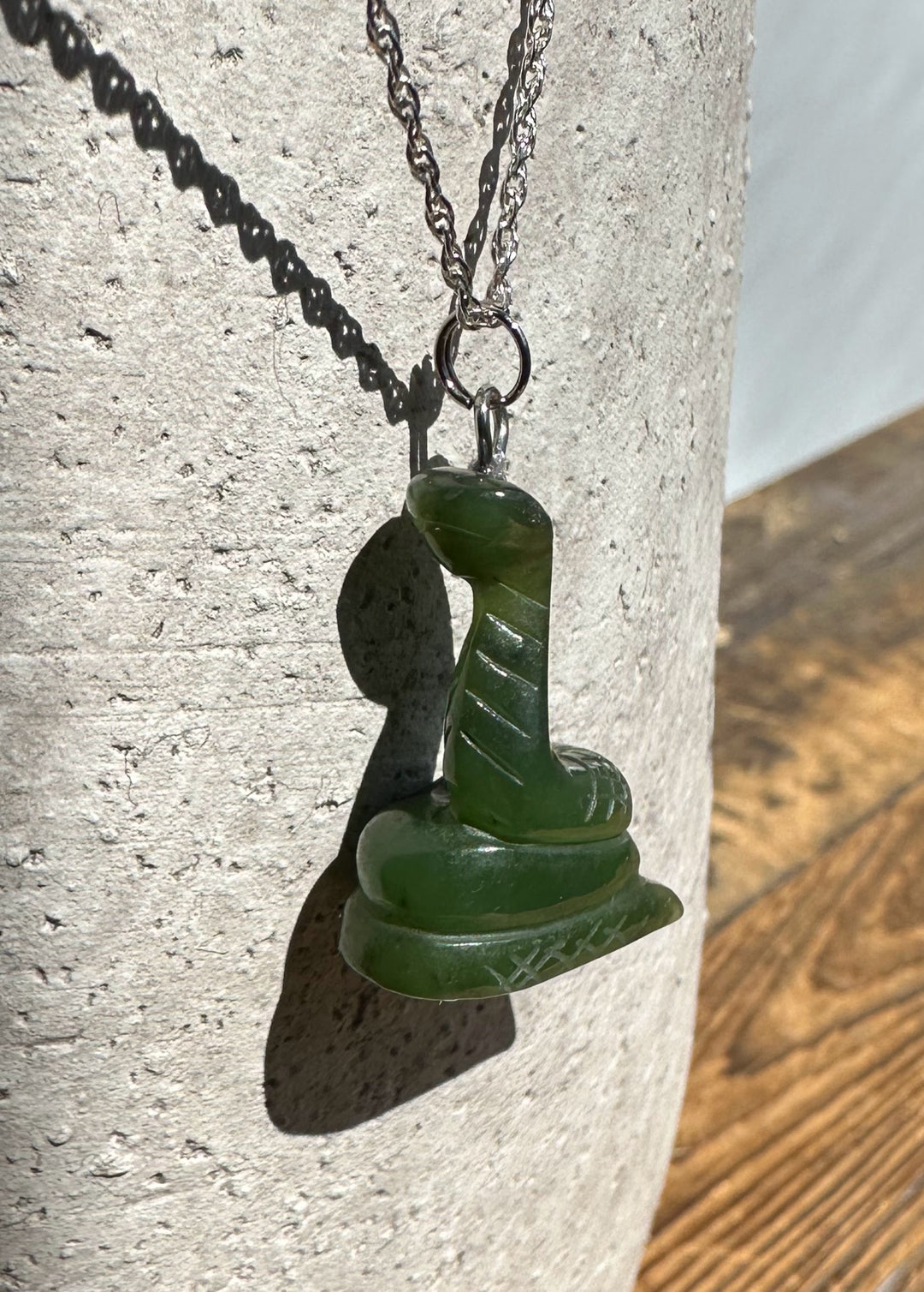 Green 1-inch jade snake pendant on a silver chain hanging against a pale concrete surface