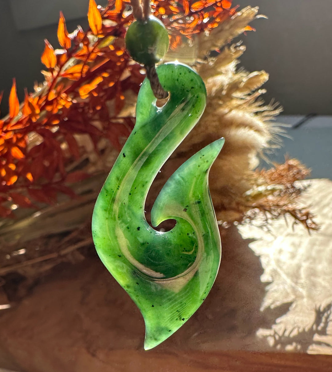 Green jade fish hook pendant with bead on cord set against orange dried-plant backdrop.