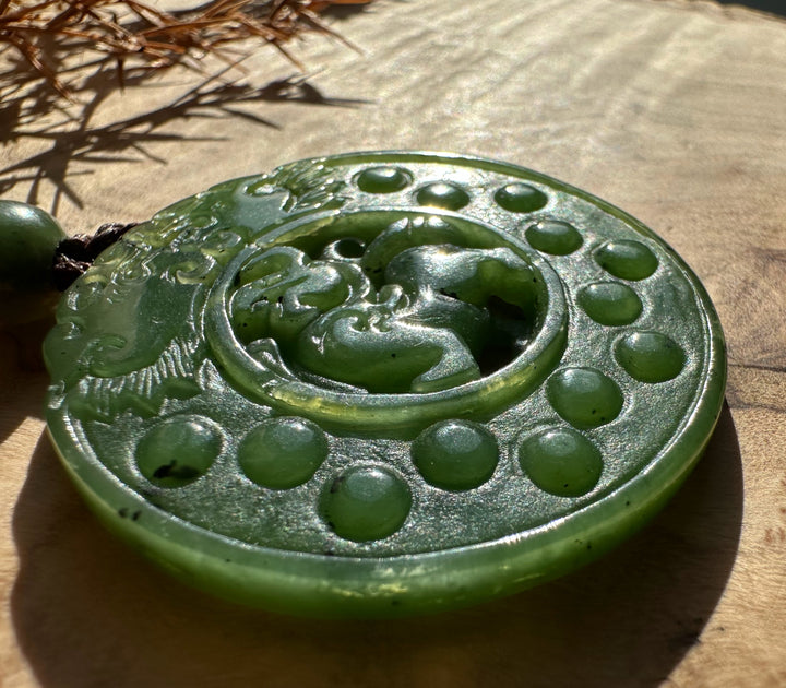 Round green jade Pixiu pendant shown from above with the carved creature and dotted border, hanging on a braided cord with a green bead.