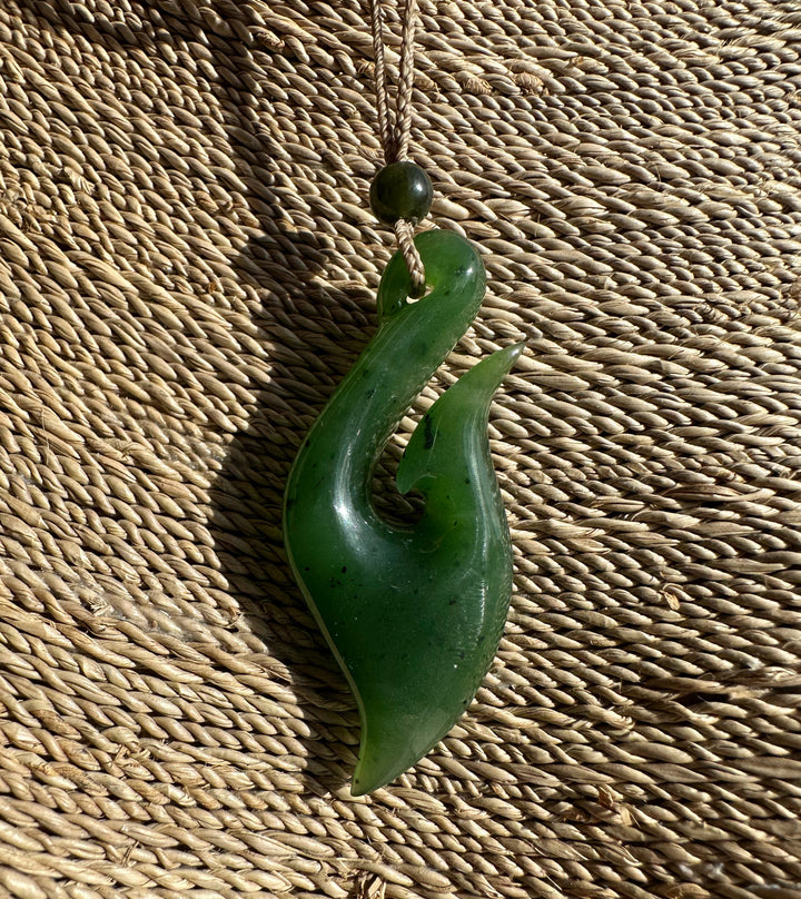 Green jade fish hook pendant on a knotted cord with a dark bead above, photographed against a woven straw backdrop