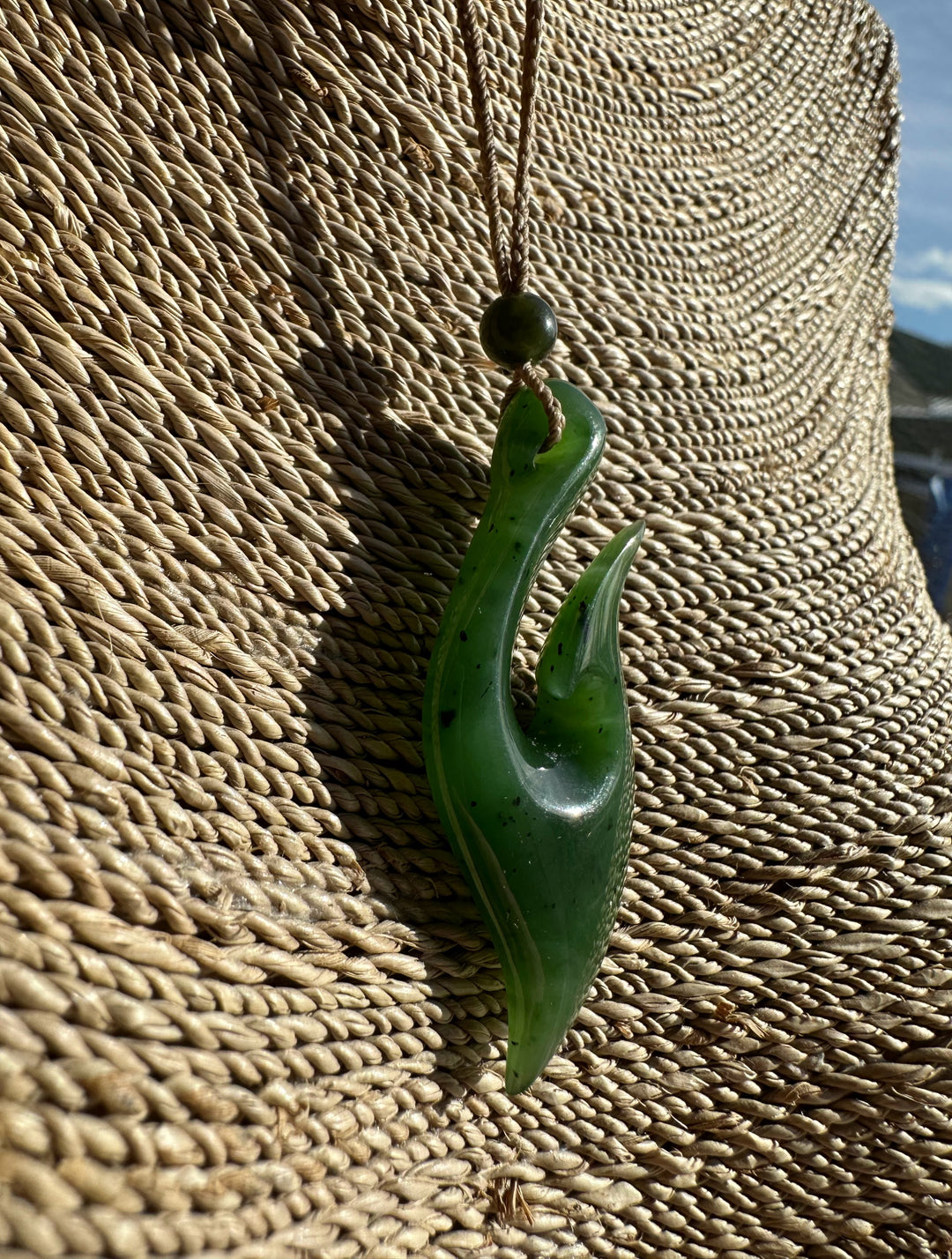 Green jade fish hook pendant with a cord and bead, hanging against a woven straw background