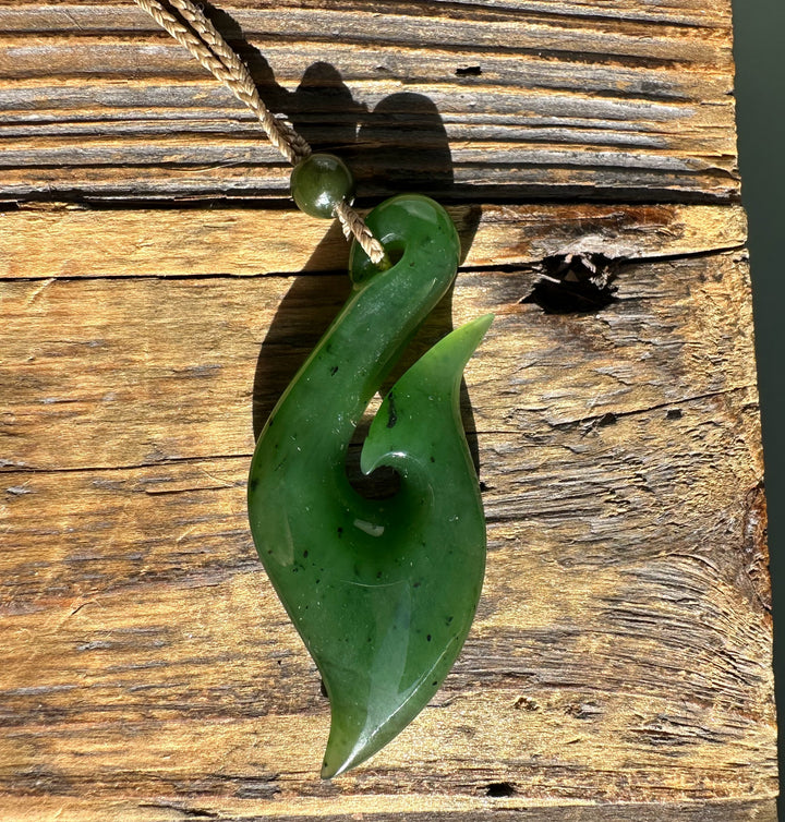 Green jade fish hook pendant on braided cord resting on weathered wood surface.