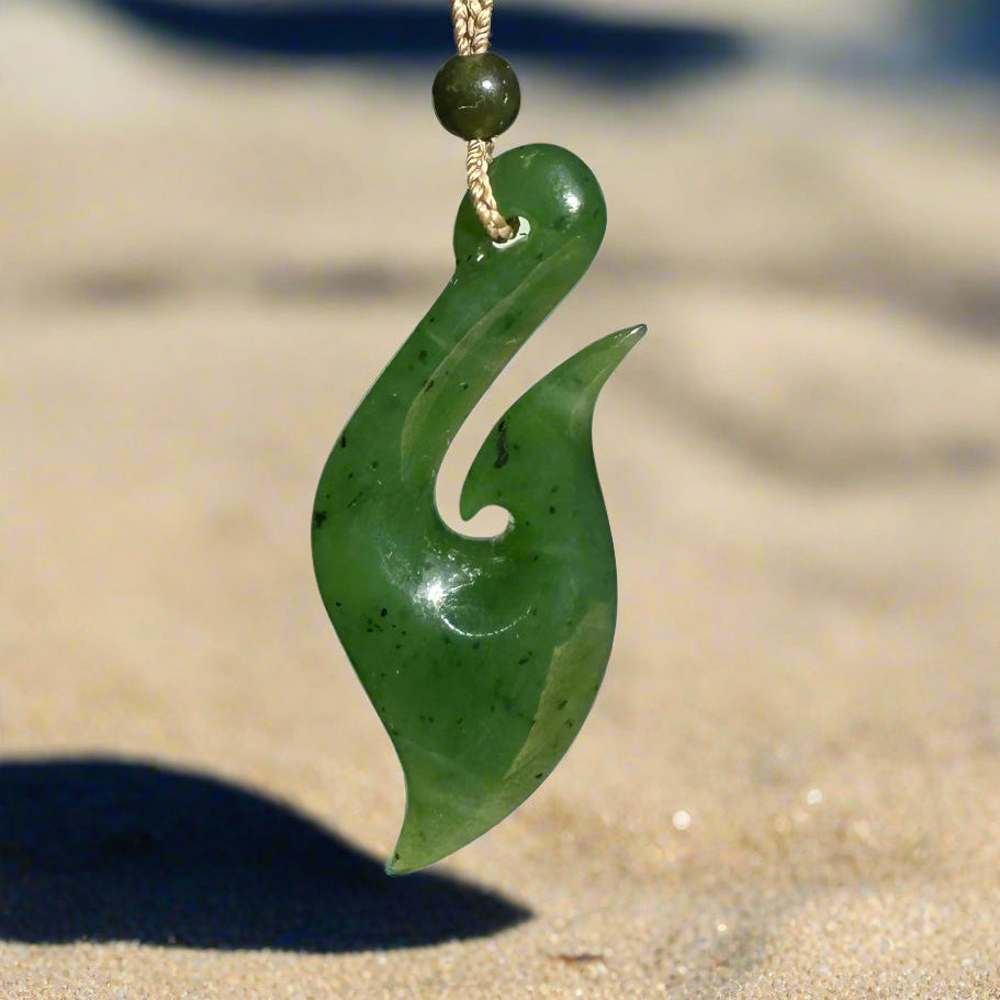 Green jade fish hook pendant, 48.5mm, hanging from a braided cord against sandy beach backdrop.