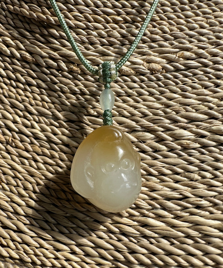Small honey white nephrite jade monkey pendant on a green cord with a decorative knot and pale beads against a woven surface