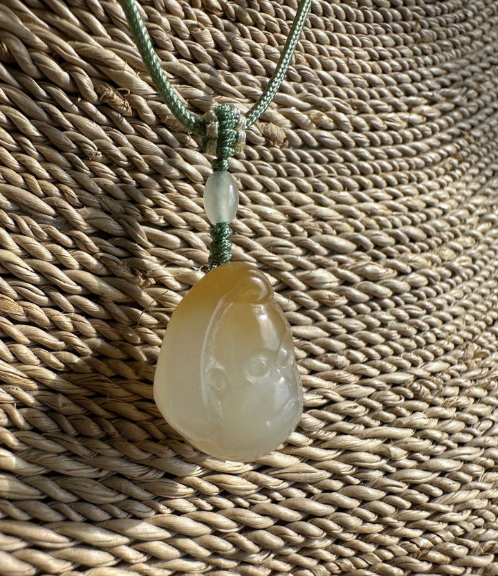 Small honey white nephrite jade monkey pendant on a green braided cord tied in a knot against a woven background