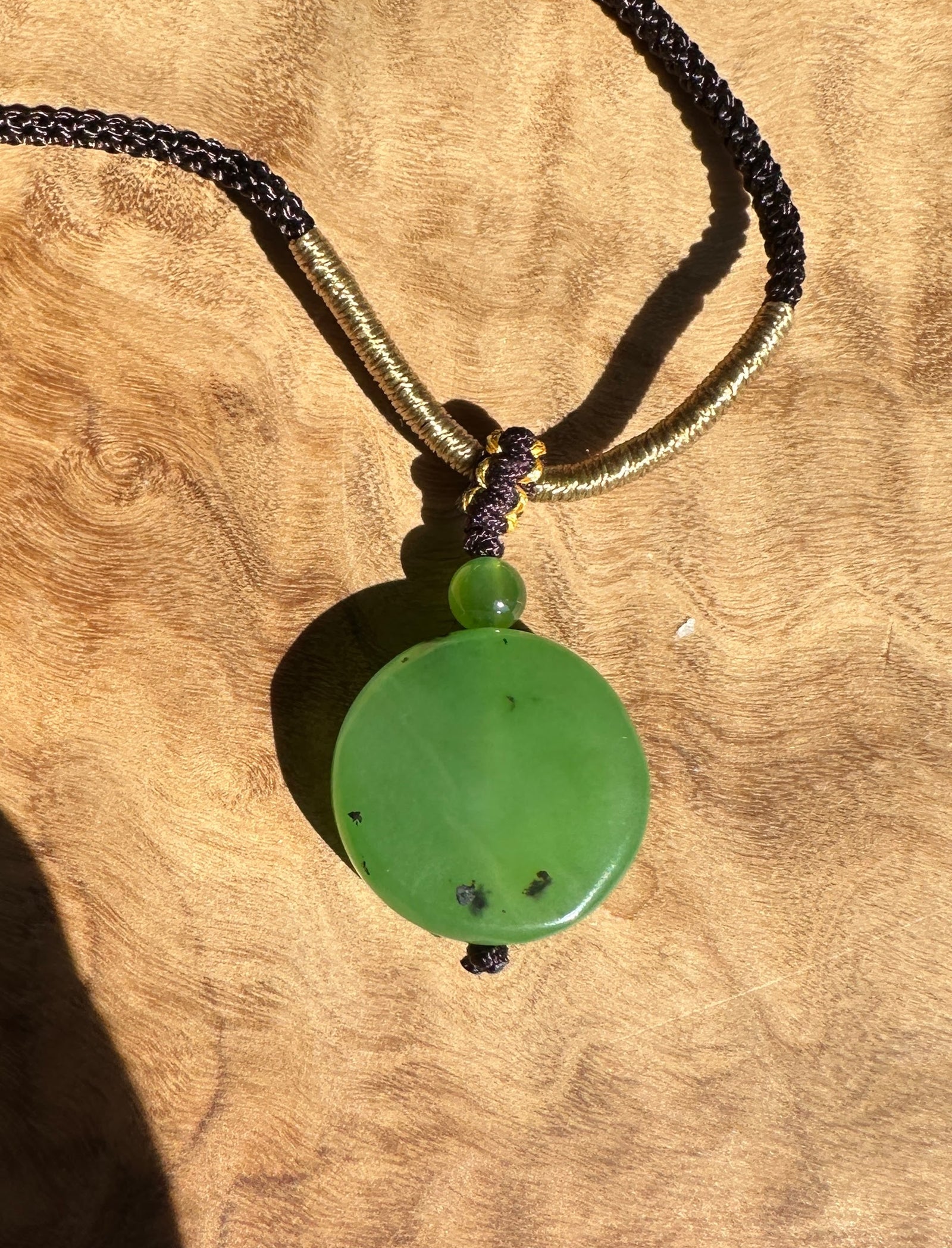 Apple green Siberian jade pendant on black and gold braided cord against a wood background.
