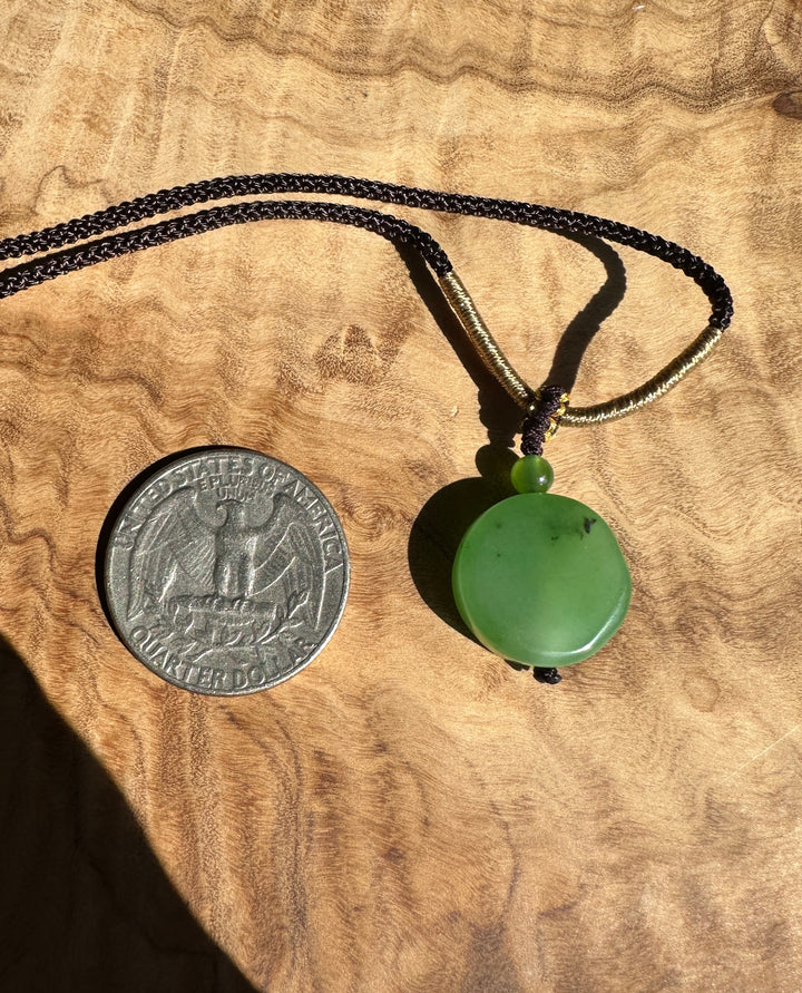 Close up of the apple green jade pendant necklace showing the gold wrapped section and dark braided cord near the pendant on wood.