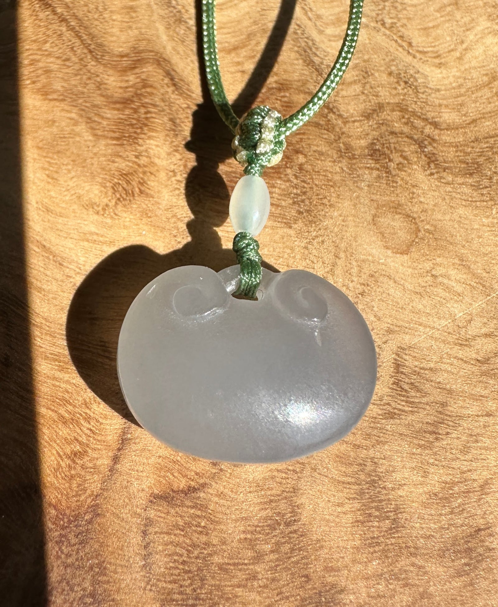 Dainty pale purple nephrite jade Ruyi pendant on a green braided cord with a white jade drop against a wood background.