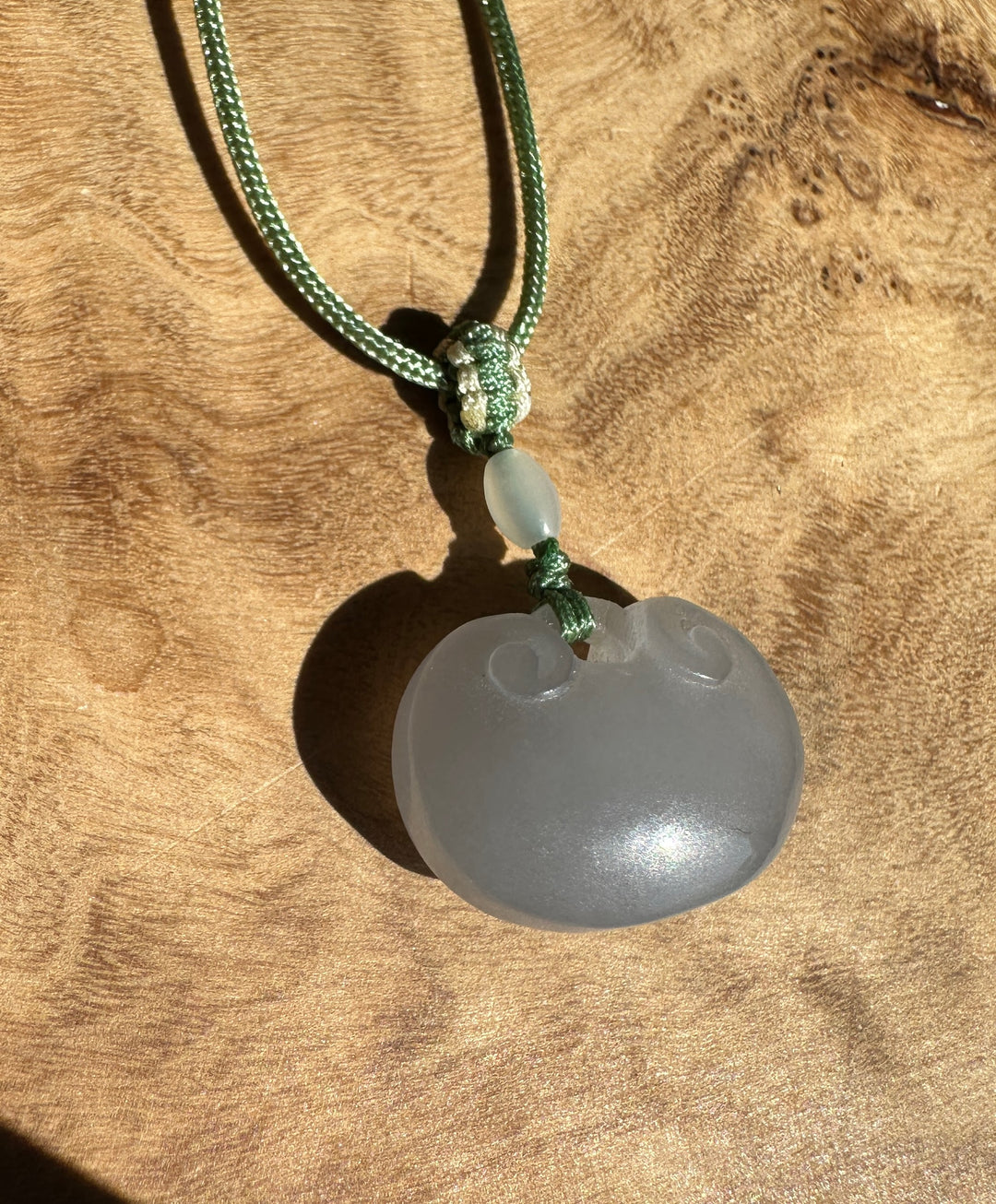 Dainty pale gray nephrite jade Ruyi pendant on a green braided cord with a knot against a wood surface