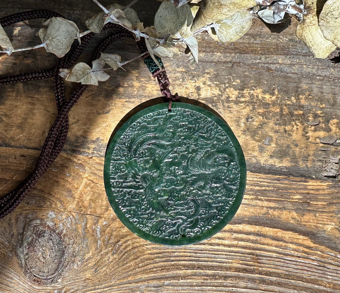 Dark green jade dragon and phoenix medallion suspended on a brown braided cord against a rustic wooden background.