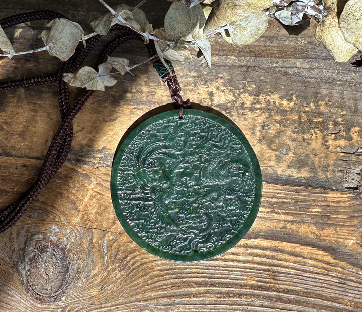 Dark green jade dragon and phoenix medallion suspended on a brown braided cord against a rustic wooden background.