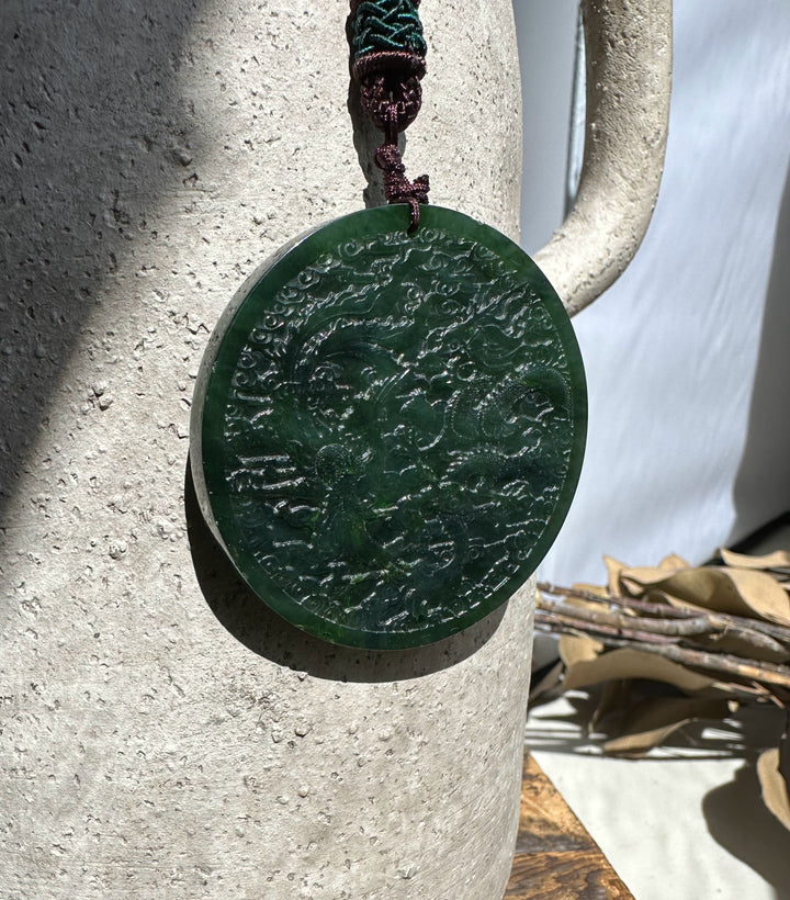 Circular jade medallion carved with dragon and phoenix, suspended from a braided cord against a stone background.