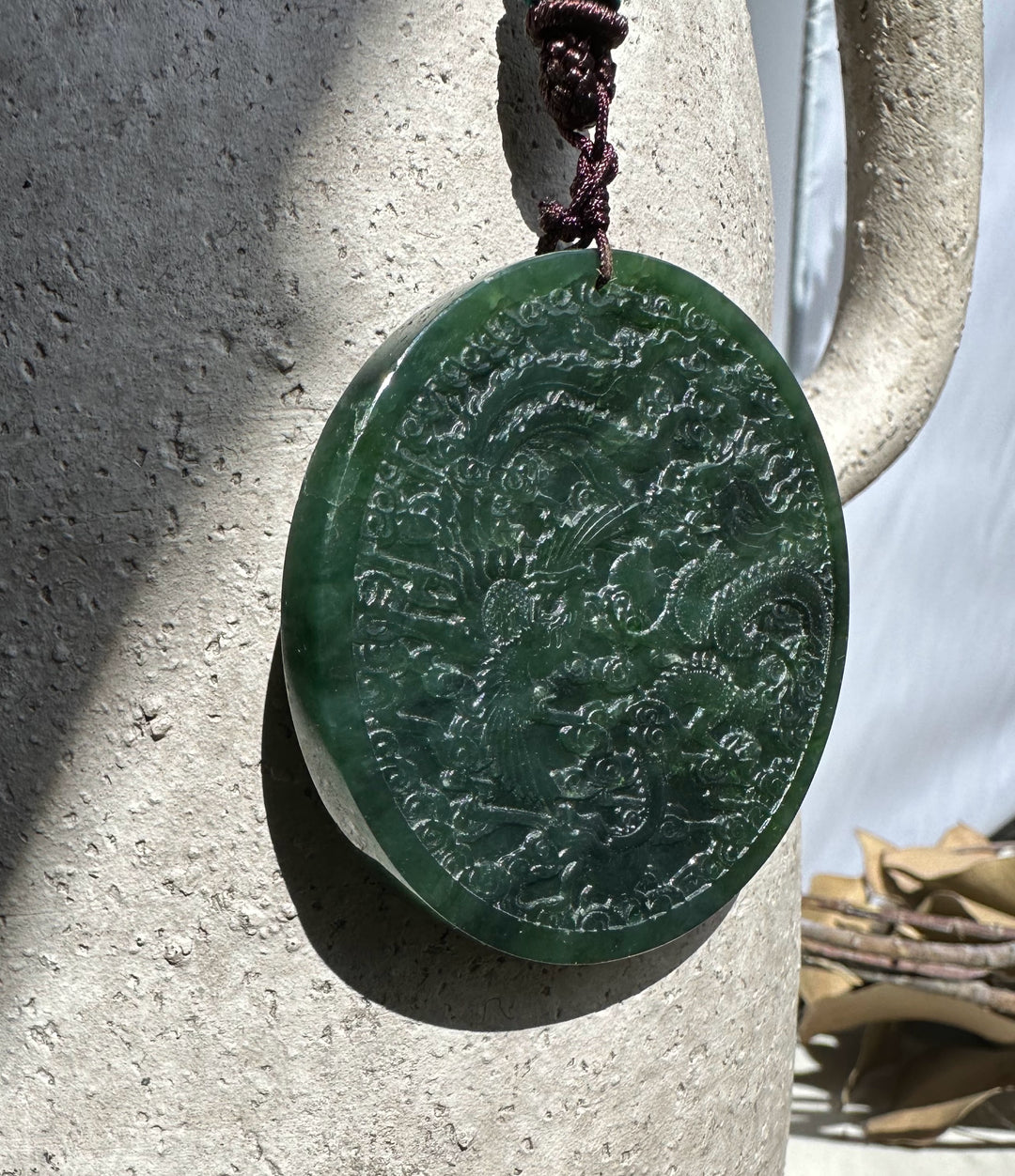 Round dark green jade medallion featuring dragon and phoenix designs hung from a brown cord beside a textured stone column.