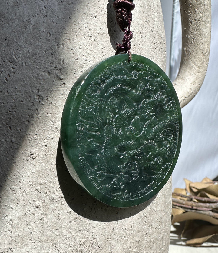 Round dark green jade medallion featuring dragon and phoenix designs hung from a brown cord beside a textured stone column.