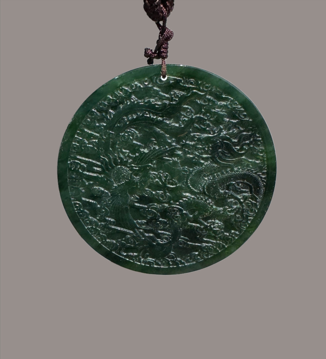 Circular dark green jade medallion with dragon and phoenix relief on a braided brown cord.