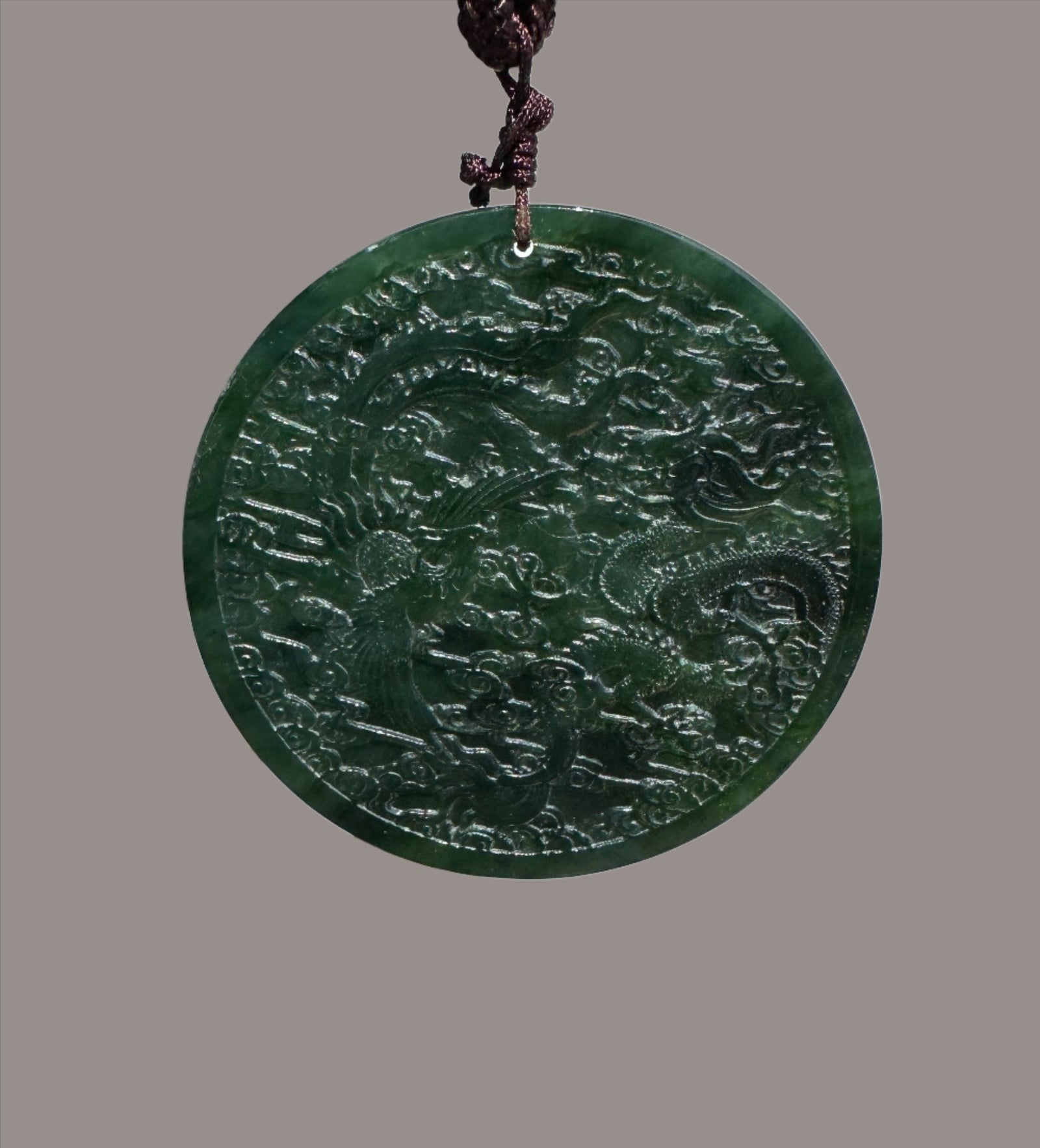 Circular dark green jade medallion with dragon and phoenix relief on a braided brown cord.