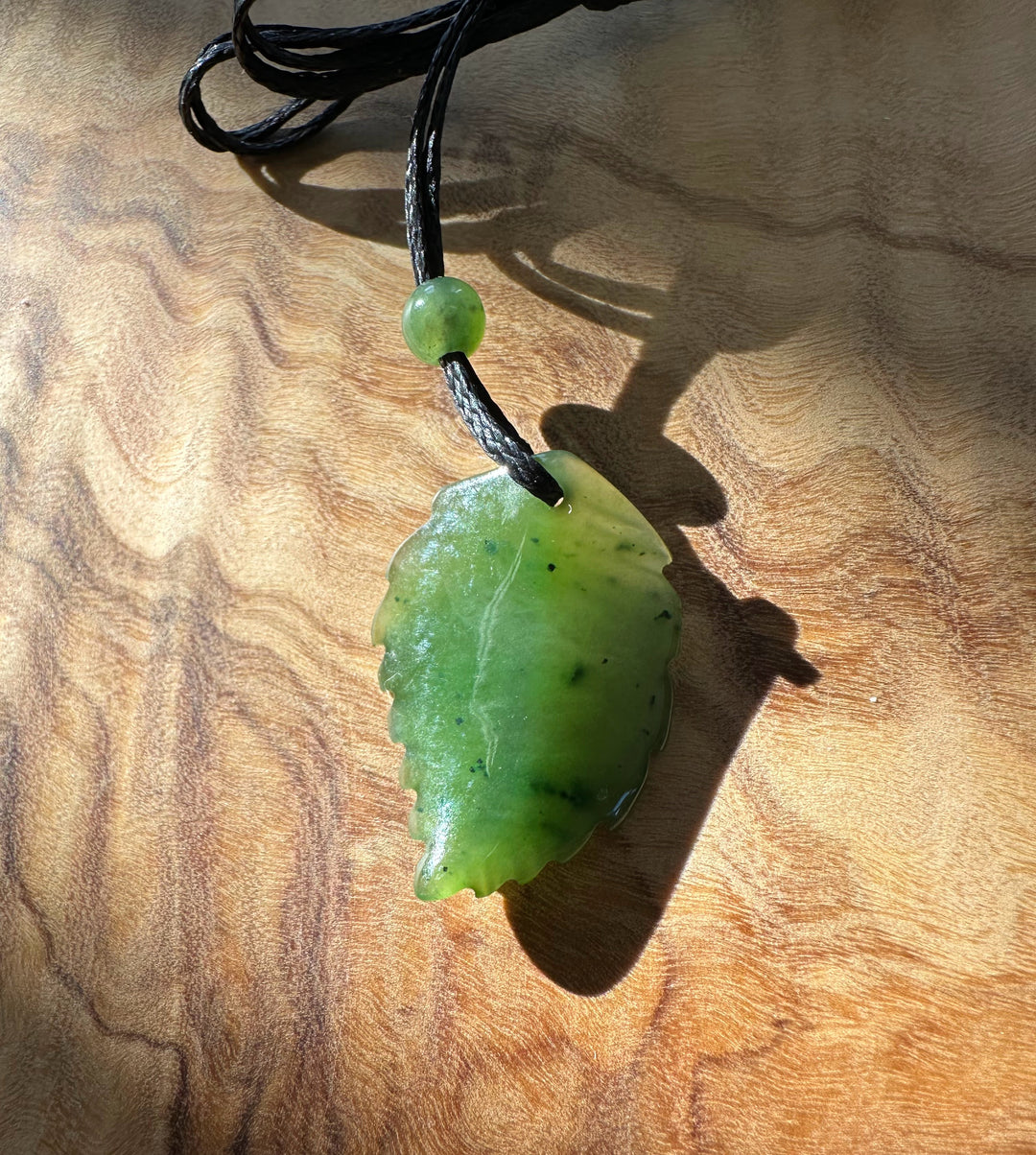 Vibrant green jade leaf pendant on a black cord with a round bead, laid on a wood surface