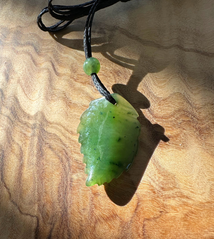Vibrant green jade leaf pendant on a black cord with a round bead, laid on a wood surface